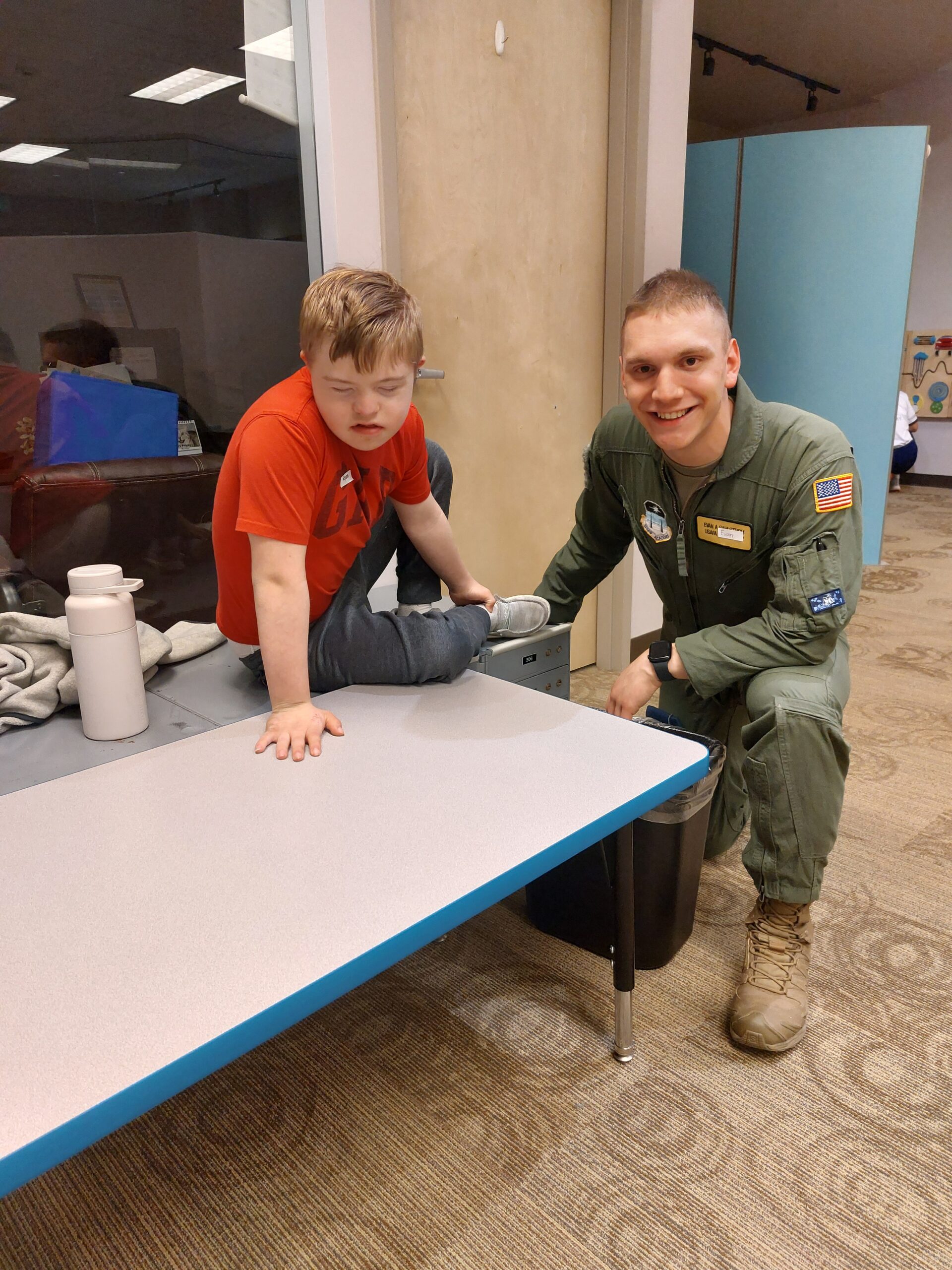 adult in a service uniform kneels next to a child crawling on a table 